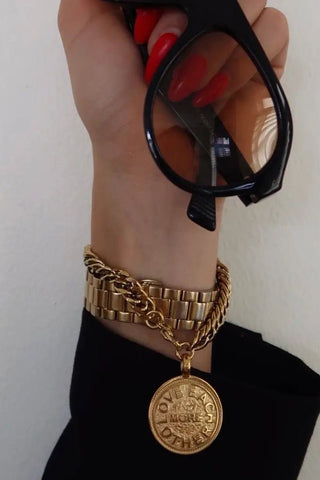 Hand wearing gold bracelets with a gold charm, holding black sunglasses against a white background